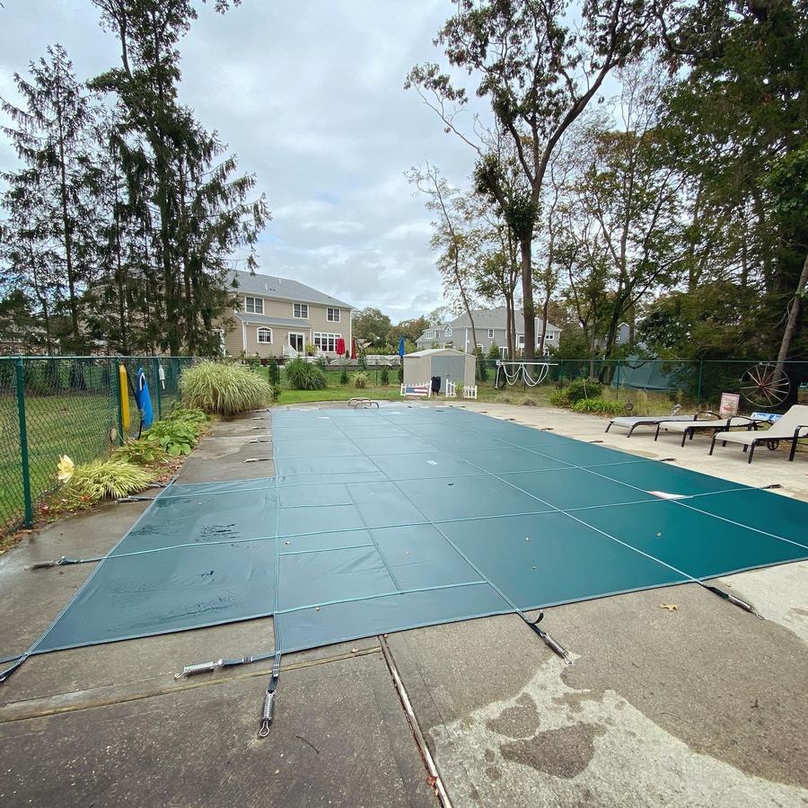 A green safety cover on a long rectangular pool in a fenced back yard.