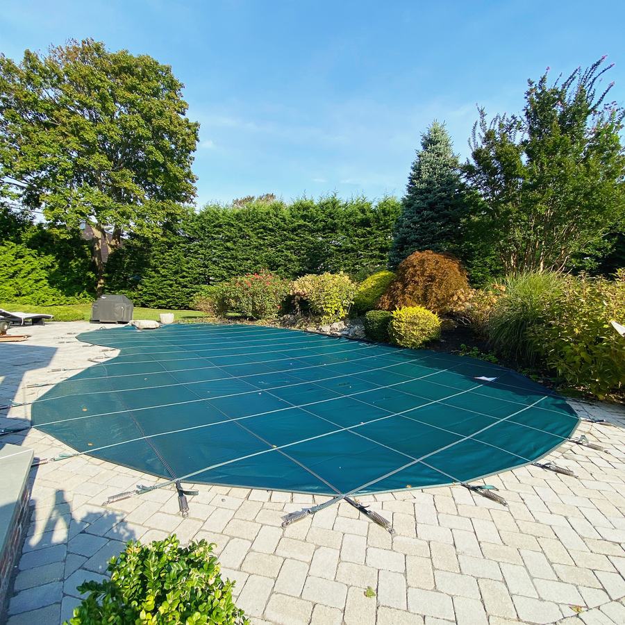 A green safety cover on a curved pool with a paver deck and flowering shrubs.
