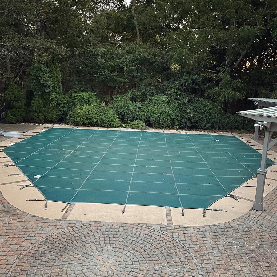 A green safety cover on an angular pool with a cobblestone border.