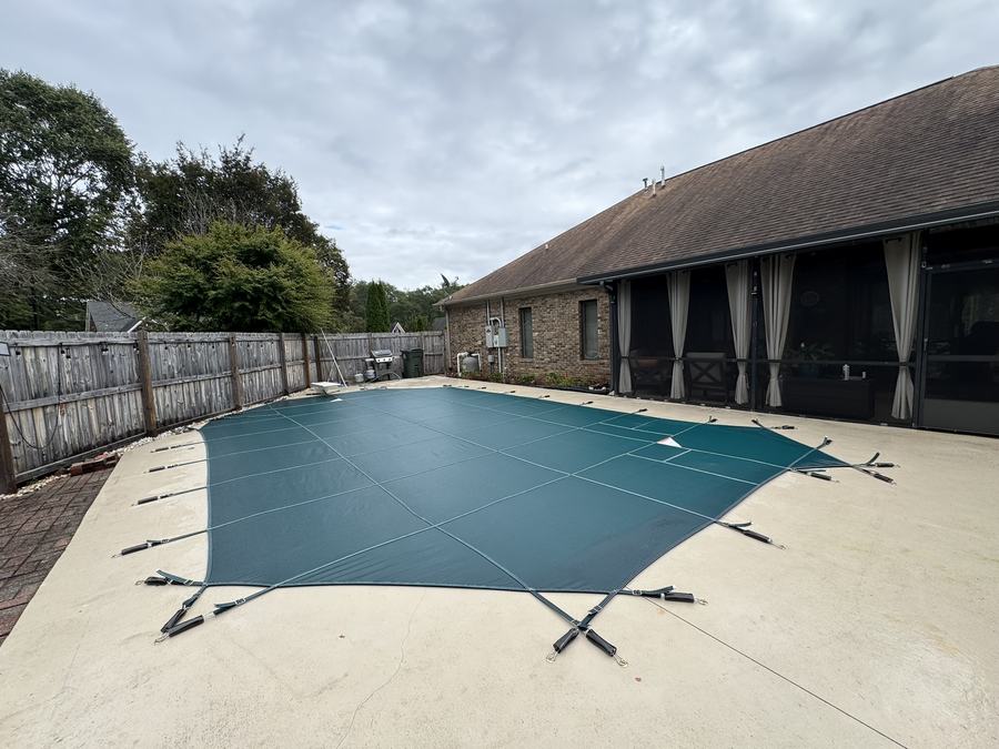A green safety cover on a rectangular pool beside a long brick home with a screened porch.