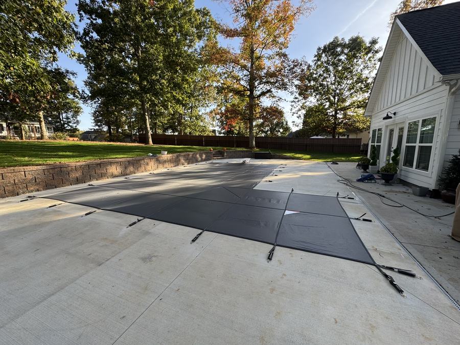 A dark safety cover on a rectangular pool beside a white farmhouse.