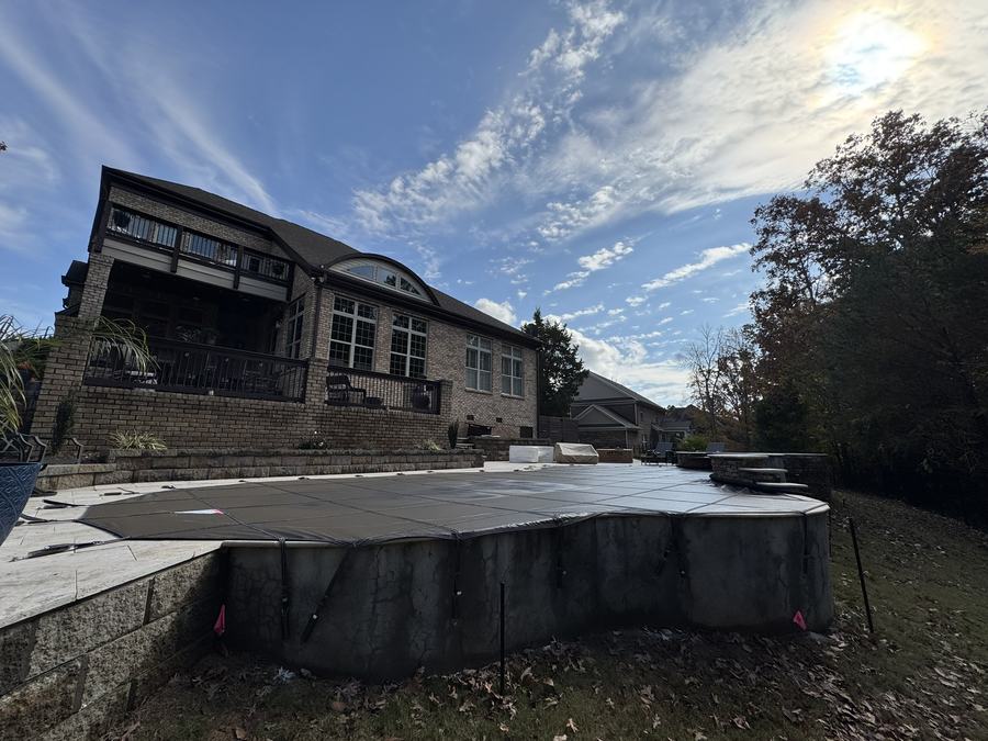 A dark safety cover on a pool with a raised wall below a large brick home.