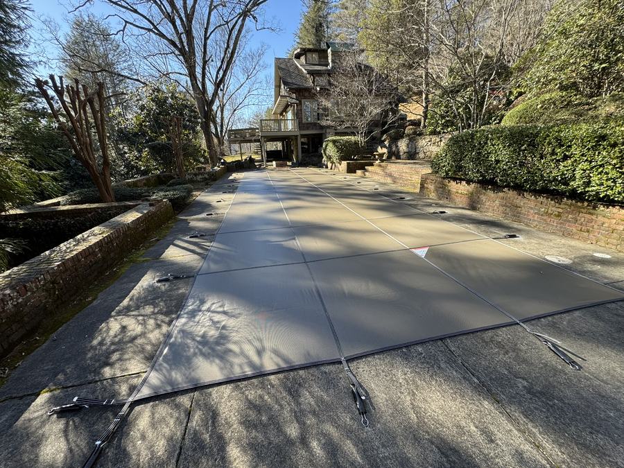 A gray safety cover on a long pool beside a stone wall in a wooded yard.