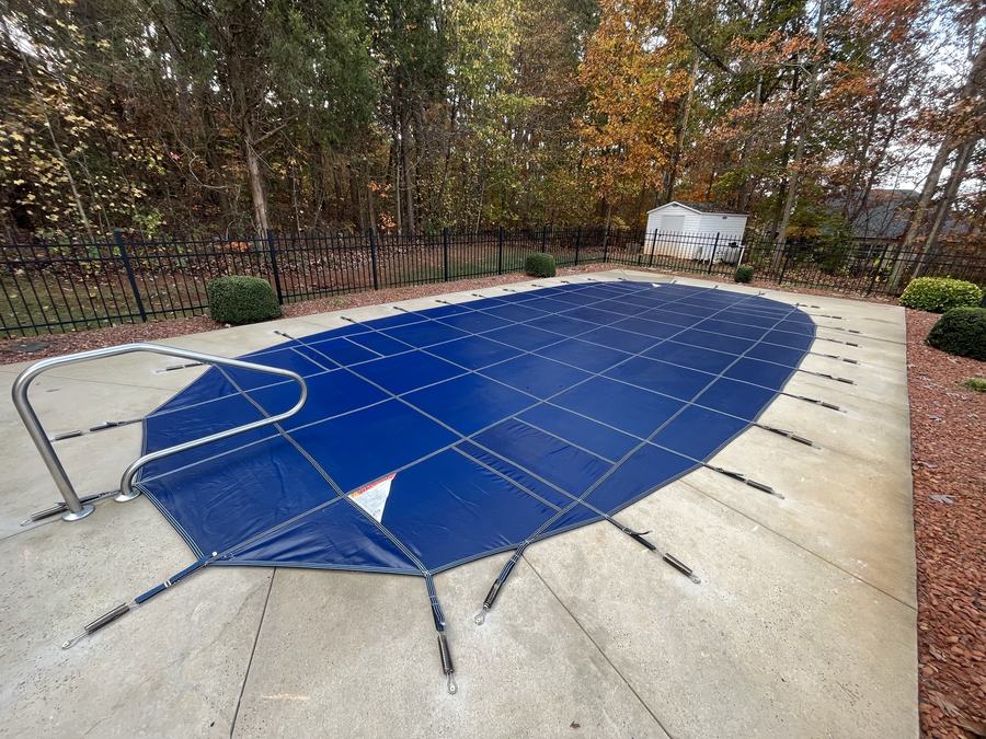 A blue safety cover on an oval pool with a handrail, surrounded by autumn trees.