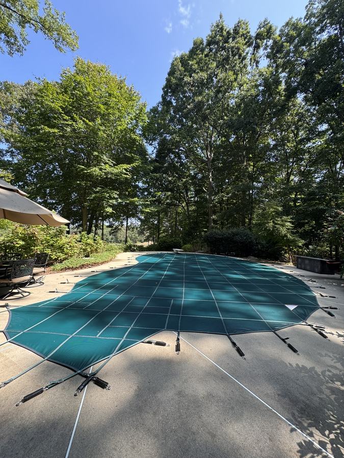 A teal safety cover on a freeform pool under tall trees.