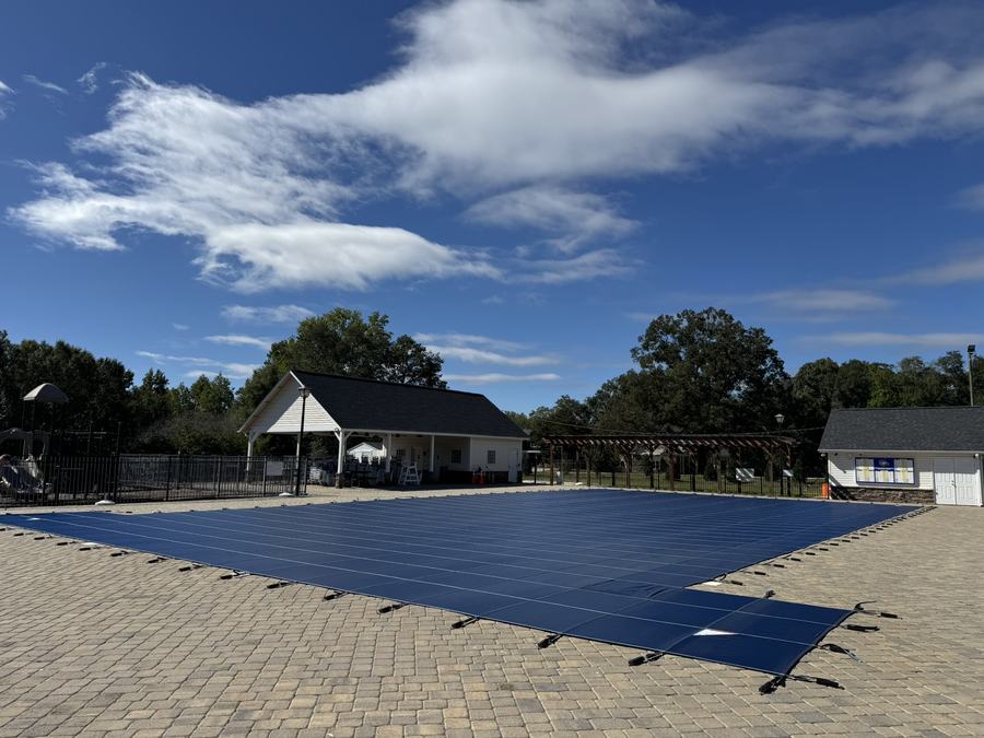 A navy safety cover on a large rectangular pool with a paver deck under a wide blue sky.