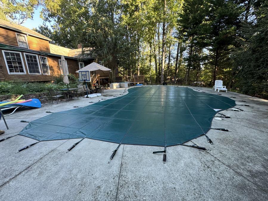 A green safety cover on a freeform pool behind a wood-sided home.