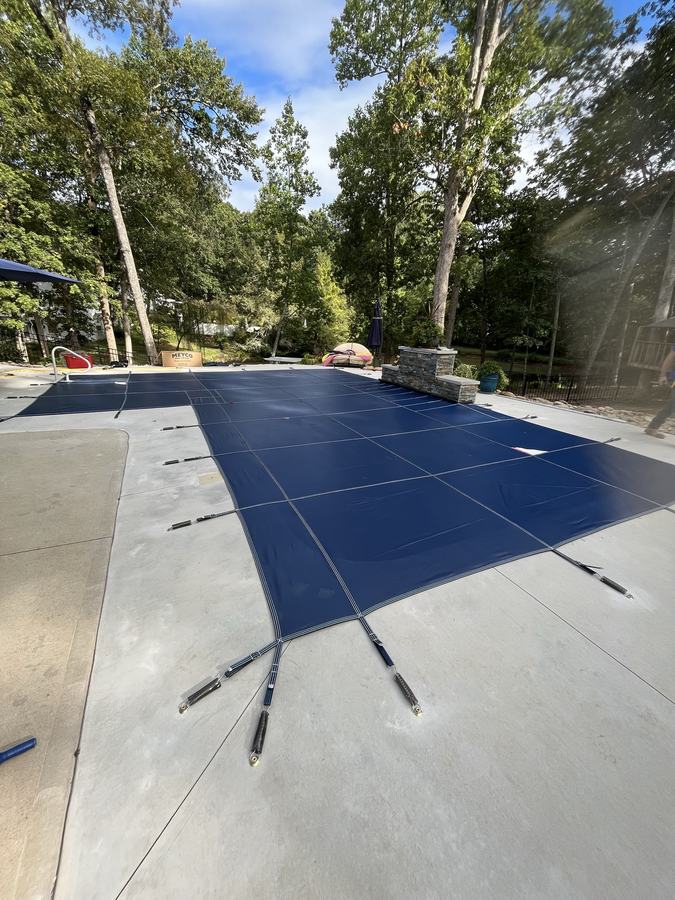 A blue safety cover on a rectangular pool with a concrete deck and tall trees.