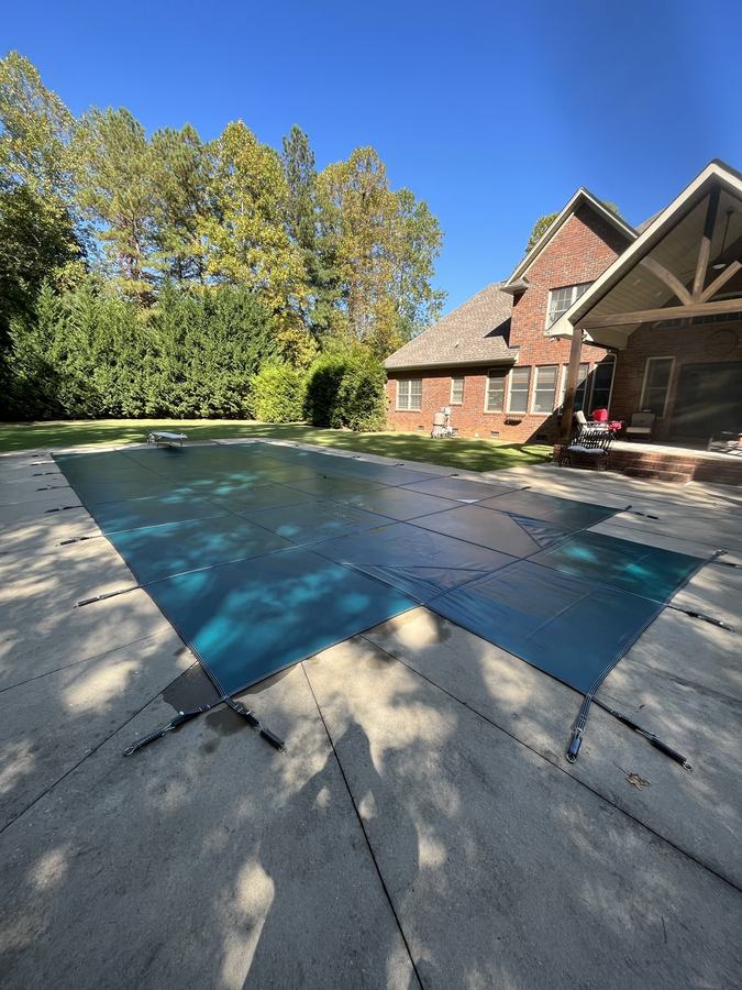 A teal safety cover on a rectangular pool beside a brick home.