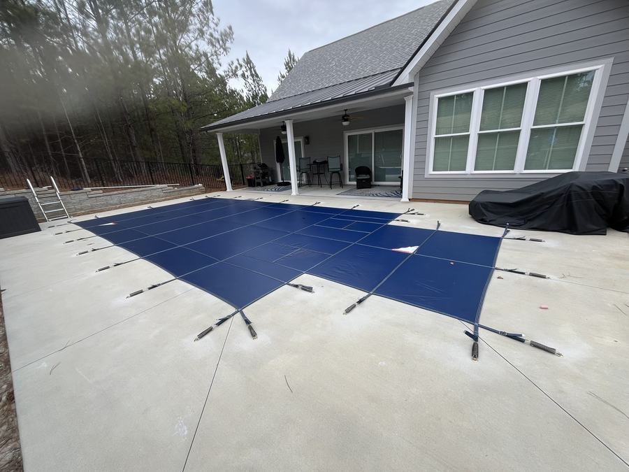 A blue safety cover on a rectangular pool behind a gray house with a covered patio.