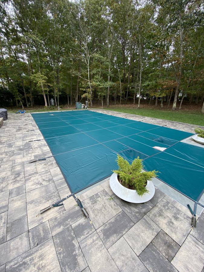 A teal safety cover on a rectangular pool with a paver deck and a potted plant.