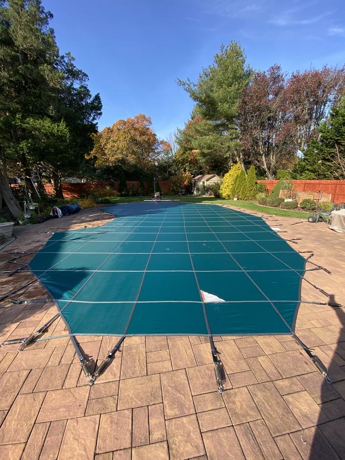 A teal safety cover on a rectangular pool with a red brick paver deck.