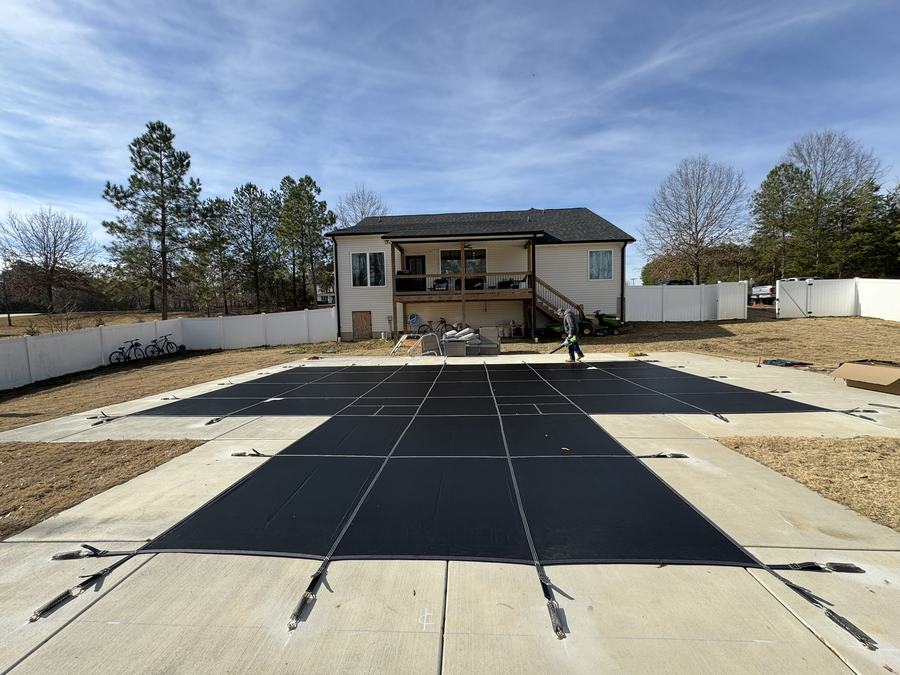 A black safety cover on a long rectangular pool behind a brick home.