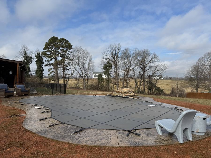 A gray safety cover on a large freeform pool beside open fields.