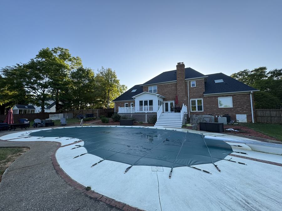 A blue safety cover on a freeform pool behind a large brick home.